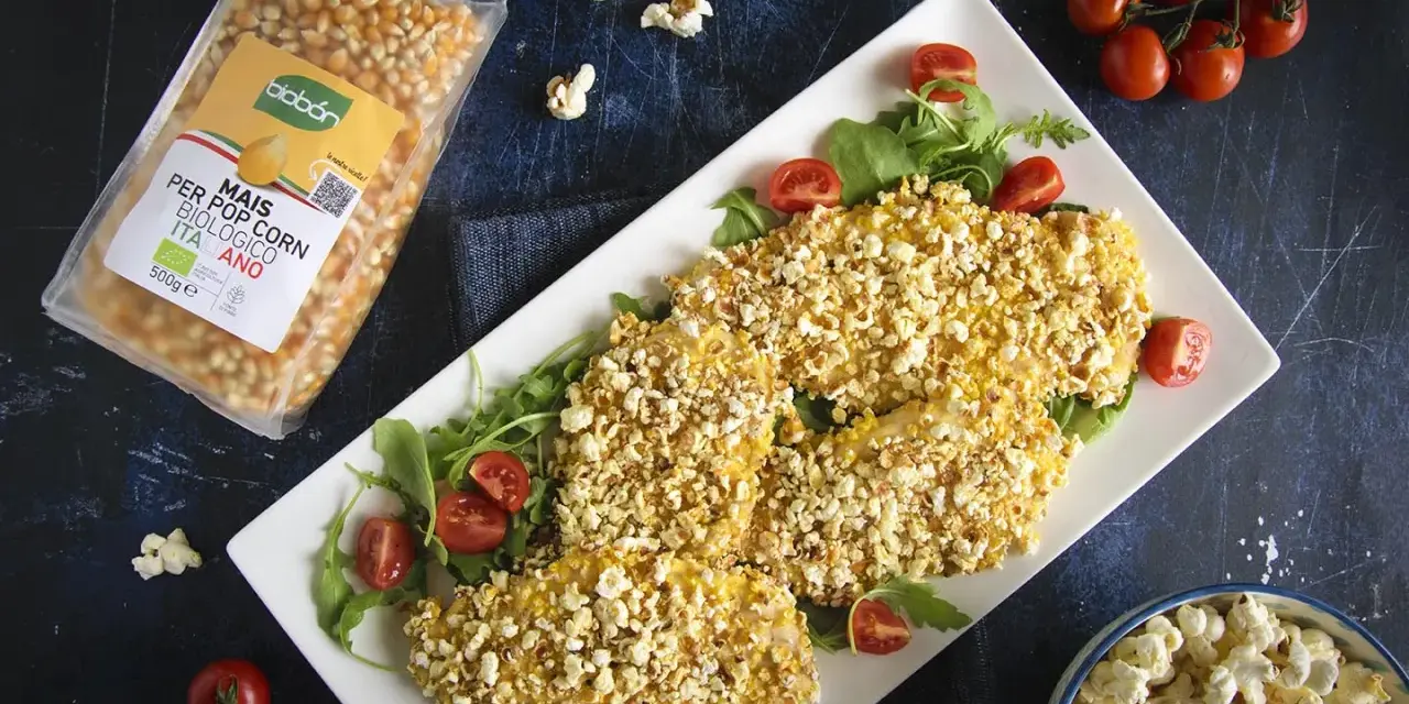 Petto di pollo impanato con **pannocchia pop corn**, servito su un letto di rucola e pomodorini. Accanto, una busta di mais per pop corn e una ciotola di pop corn.