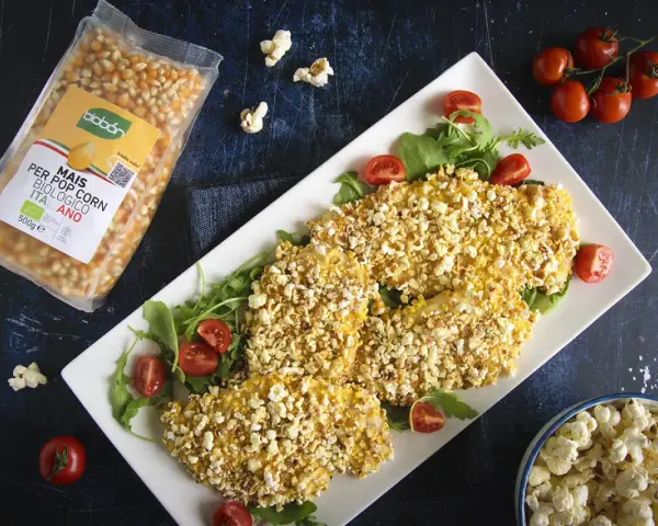 Petto di pollo impanato con **pannocchia pop corn**, servito su un letto di rucola e pomodorini. Accanto, una busta di mais per pop corn e una ciotola di pop corn.