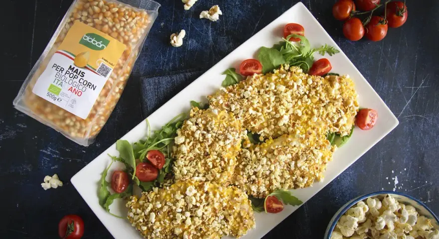 Petto di pollo impanato con **pannocchia pop corn**, servito su un letto di rucola e pomodorini. Accanto, una busta di mais per pop corn e una ciotola di pop corn.