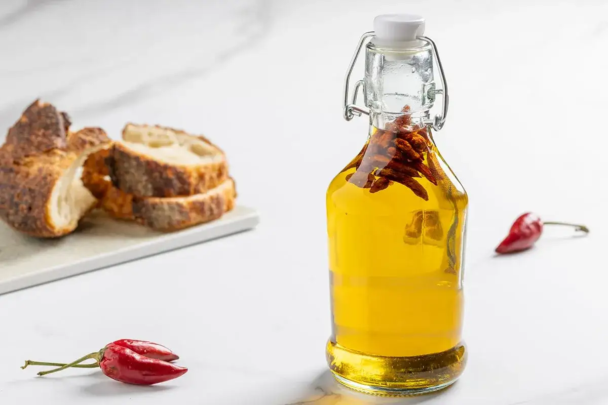 Olio dorato in bottiglia con peperoncino piccante, accanto a fette di pane rustico e un altro peperoncino rosso.